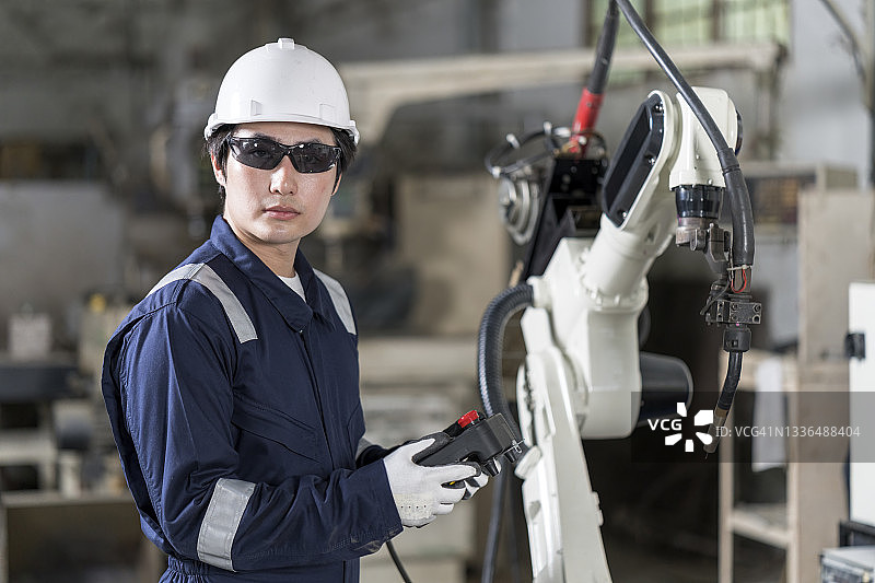 男工业工程师手拿数字控制器站在金属工厂的机器人焊接机旁边。图片素材