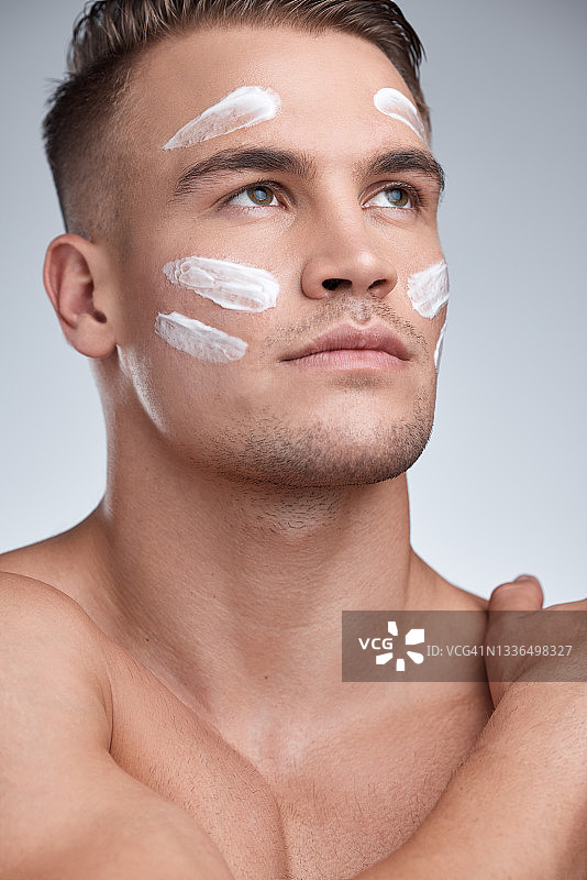 一个有魅力的年轻男子在灰色背景下往脸上涂抹润肤霜的短发特写图片素材