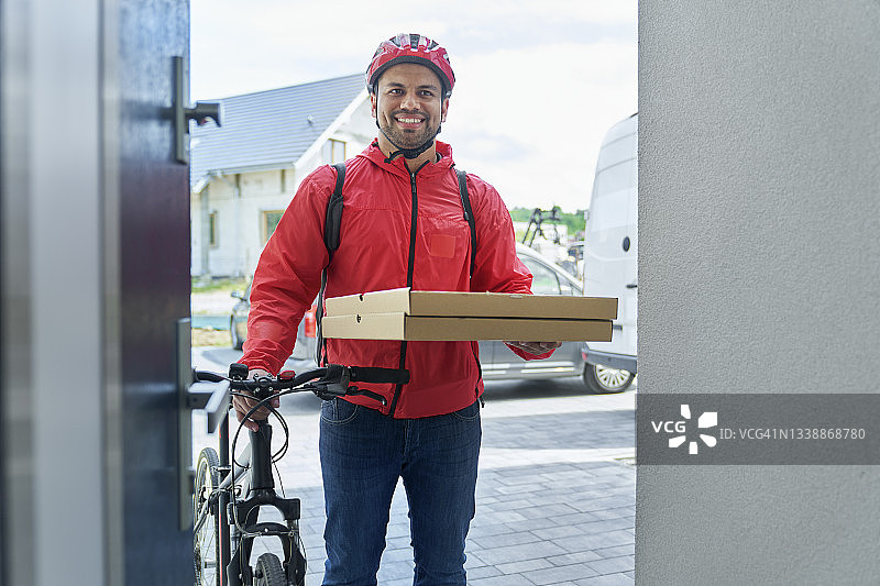 微笑的送餐员拿着披萨盒，骑着自行车站在门口图片素材