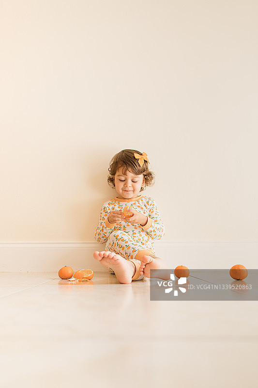 2021年9月，两岁的小女孩坐在地板上玩和吃橘子，穿着橙色睡衣和橙色的头发蝴蝶结在家里图片素材