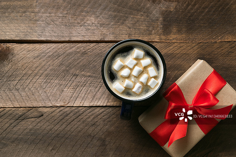 准备一个家庭节日的概念，平安夜，情人节或生日快乐。圣诞礼物包在鲜红的丝带里，背景是一张木桌。一杯热咖啡或加棉花糖的可可。图片素材