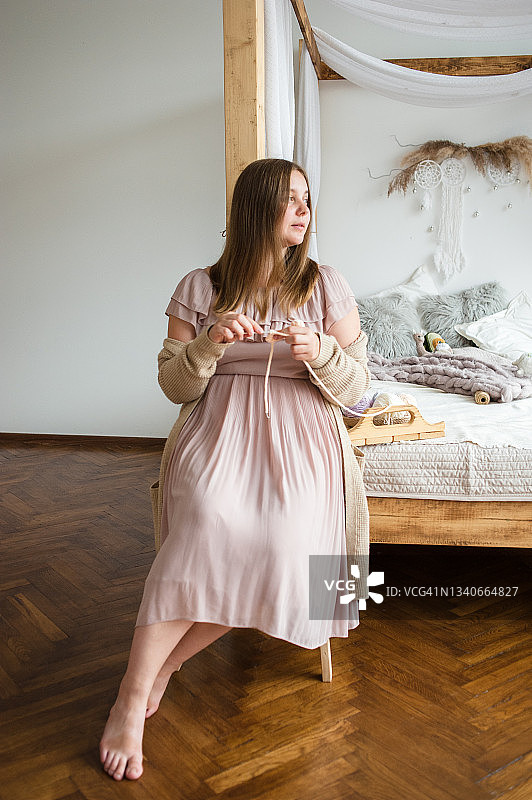 美丽的年轻女子，一个编织玩具的大师，坐在波西米亚风格的卧室靠在顶篷床上，编织着，看着远方。在她身后的床上放着她亲手制作的玩具，还有木盘上篮子里的一缕缕羊毛。图片素材