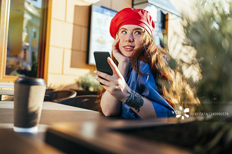 一名戴着贝雷帽的年轻女子在路边咖啡馆拿着手机做着白日梦图片素材
