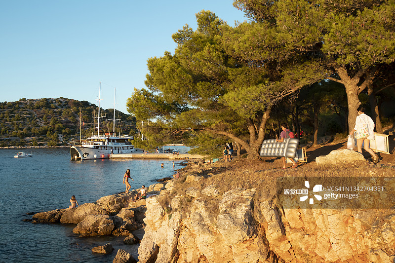 克罗地亚，默特岛，岩石沙滩旁的松树，沙滩上的人们带着沙滩配件，在夏日的夕阳下停泊着小船图片素材