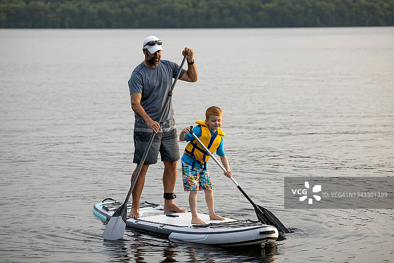 在一个温暖阳光的暑假里，父亲和儿子在安静的湖上站着冲浪图片素材