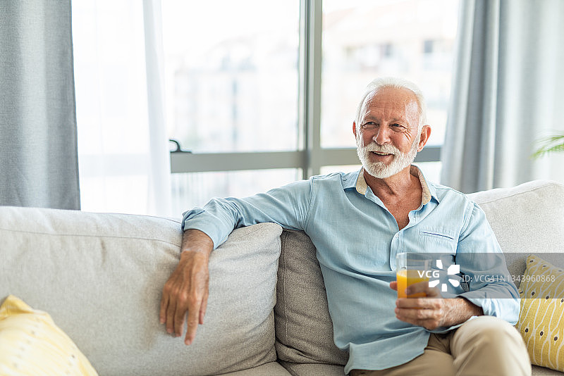 快乐的老人坐在家里的沙发上喝着橙汁。图片素材