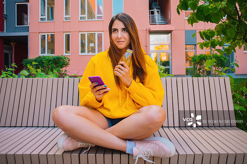 一位身穿黄色连帽衫的年轻迷人女子坐在长椅上使用智能手机图片素材