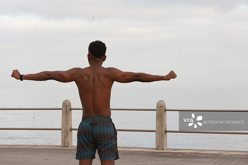 一个强壮的赤膊年轻男子在户外伸展双臂，从木板路或散步道上俯瞰自然的海洋景色图片素材