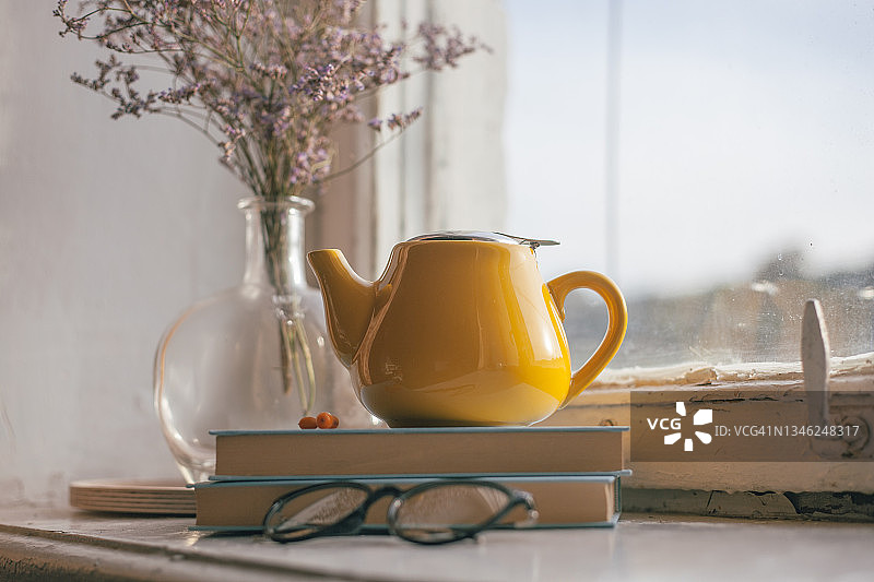 家庭内部的细节。黄色的茶壶，书籍和木制窗台上紫色花朵的古董玻璃花瓶。图片素材