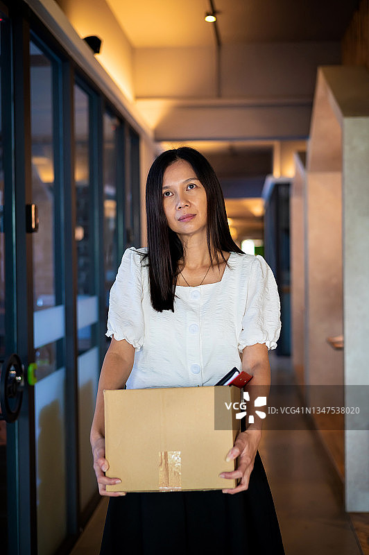 一个紧张的亚洲女人拿着一个装着个人物品的纸板箱站在办公室走廊上。图片素材