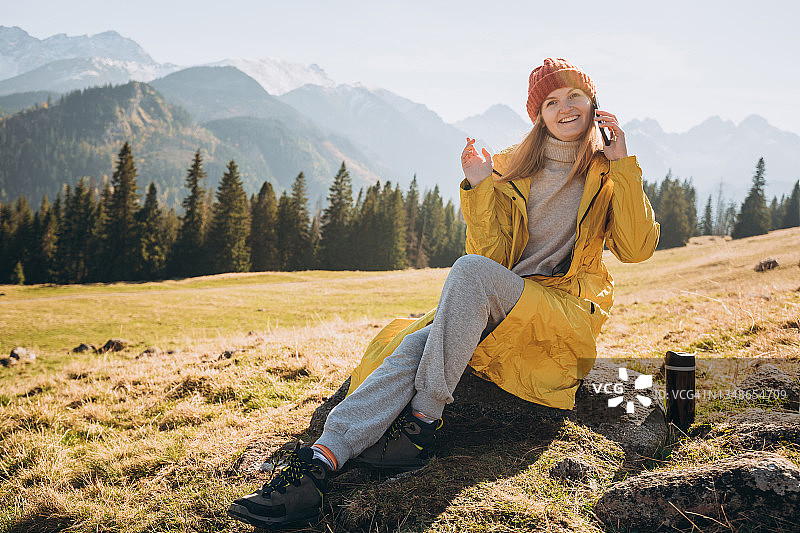 年轻的金发女子坐在山里，用智能手机聊天。自由，快乐，旅游和度假的概念，户外活动图片素材