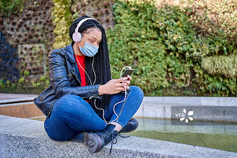 一名非裔美国妇女的肖像戴着口罩，拿着手机在城市的街道上听音乐。头发被编成了脏辫。新常态的概念图片素材