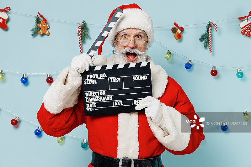 圣诞老人戴着圣诞帽，身穿红色套装，手持经典的黑色电影制作隔板，孤立在素色背景的摄影棚。祝2022年新年快乐，庆祝圣诞佳节快乐。图片素材