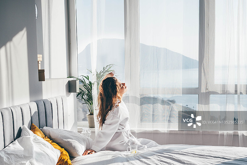 睡醒后，身穿白色浴衣的女子坐在白色大窗旁，在床上伸展身体。图片素材
