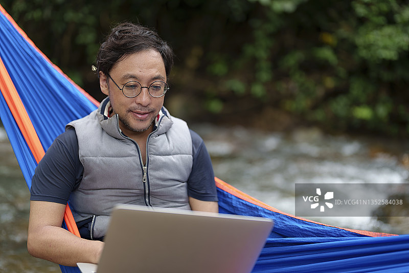 在河边的森林里，一名亚洲日本男子坐在吊床上使用笔记本电脑。以现代科技为生活方式的工作和旅游理念。图片素材