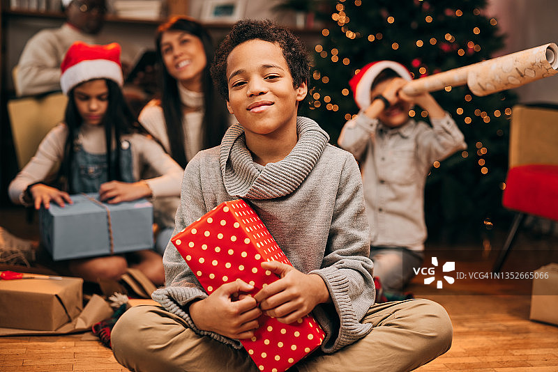 一个快乐的混血男孩在圣诞节坐在家里的地板上，手里拿着一份礼物。在模糊的背景中，他的多种族家庭在一起玩耍。图片素材