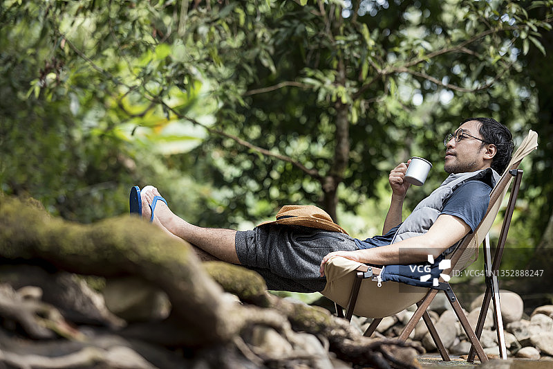 自然的声音能帮助你放松。日本男人在露营时坐在椅子上喝热咖啡，听着大自然的声音来放松。图片素材
