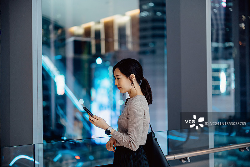 专业年轻的亚洲女商人站在当代办公空间的窗口，看着手中的智能手机。夜晚，在市中心明亮的城市天际线的映衬下。忙碌的生意图片素材