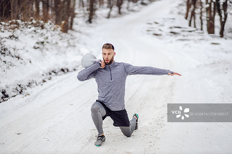 健康的运动员在树林的雪道上举壶铃做箭步。冬季健身，健康生活，健美图片素材