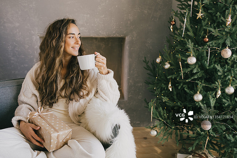 年轻女子喝热咖啡或可可坐在沙发上舒适的房间与圣诞树。冬天的假期。图片素材