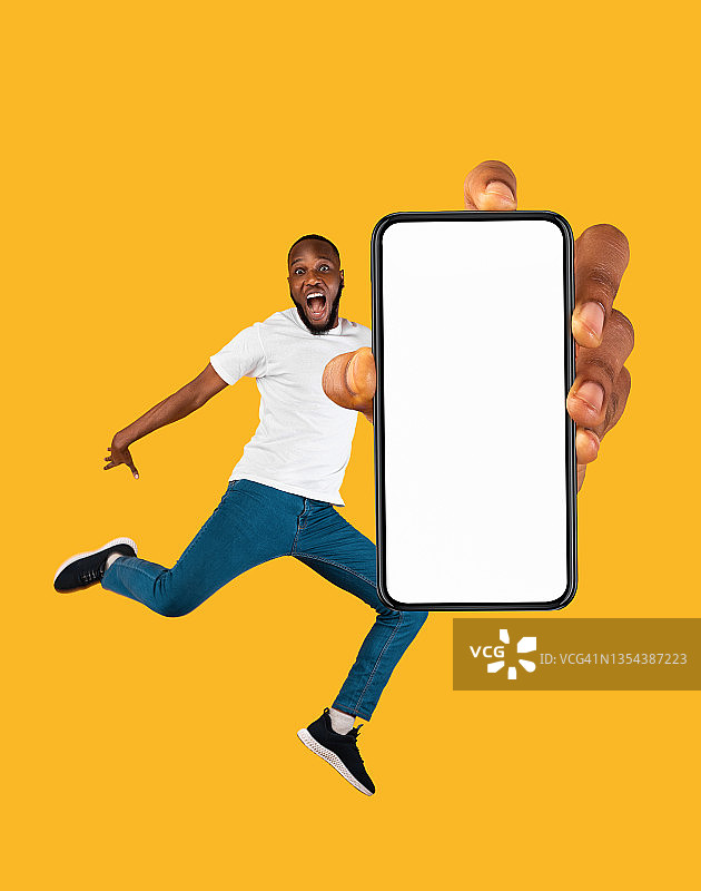 欣喜若狂的年轻黑人跳了起来，展示了白色屏幕和橙色背景的智能手机模型图片素材