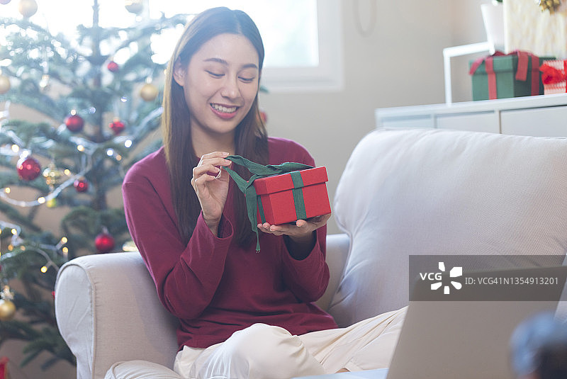 亚洲妇女收到圣诞礼物和打开盒子，坐在舒适的客厅沙发上。图片素材
