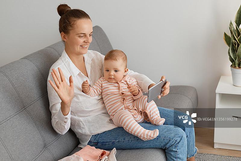 快乐妈妈和可爱宝宝用智能手机直播或视频通话的室内镜头，女人对着手机摄像头挥手问好。图片素材