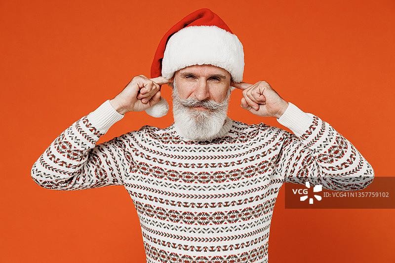 老惊慌害怕伤心大胡子圣诞老人50多岁戴圣诞帽毛衣摆姿势闭上眼睛捂住耳朵用手手指不想听尖叫孤立在纯橙色背景工作室图片素材