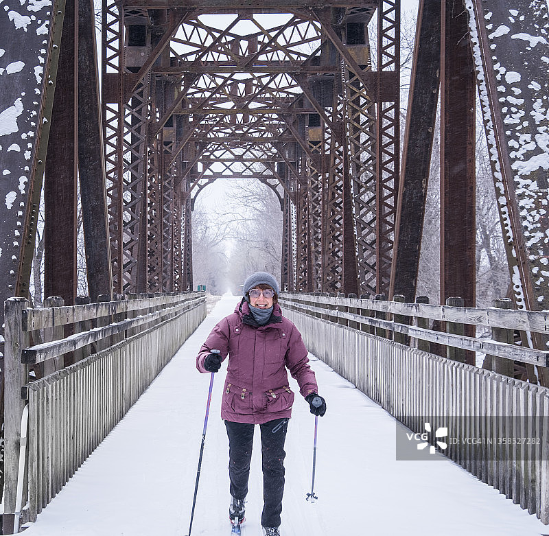 在中西部的雪天，微笑的老妇人在旧铁路桥下越野滑雪转移到国家娱乐小径图片素材