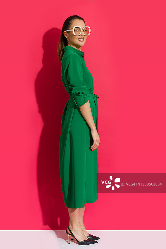 一个穿着绿色长扣裙、高跟鞋、戴着大墨镜的年轻微笑的女人靠着红色的墙站着。图片素材