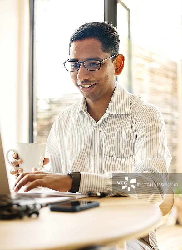 微笑的年轻商人在家里喝着咖啡，用笔记本电脑工作图片素材