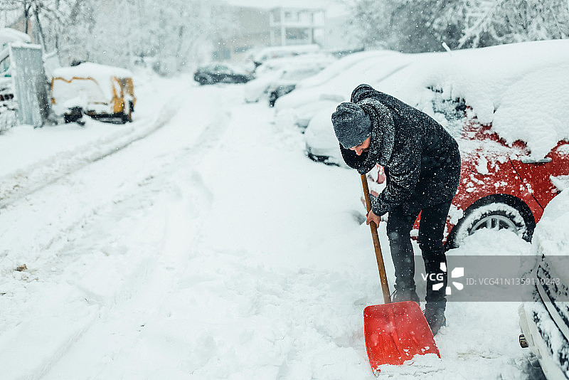 拿着雪铲的男人。积雪下的汽车和街道。图片素材