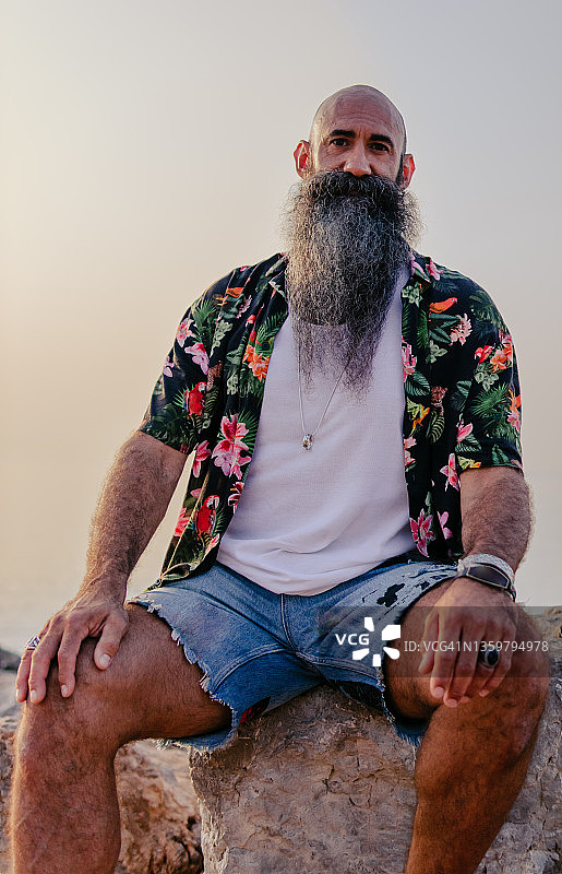 一名留着长胡子、穿着夏威夷绿色衬衫的男子坐在沙滩上的一块岩石上的肖像图片素材