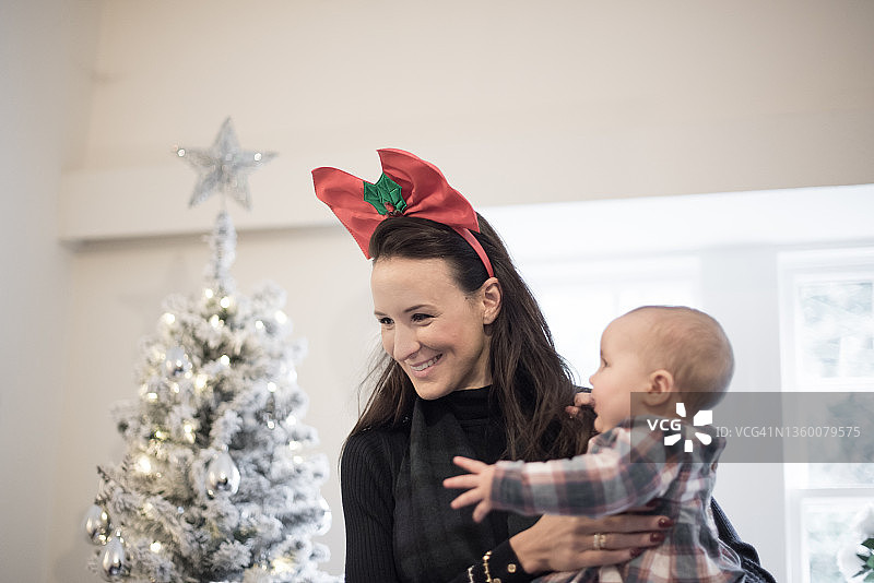 在英国苏格兰边境梅尔罗斯的一所房子里，一位年轻的母亲戴着红色的圣诞蝴蝶结头带，在窗边的一棵白色圣诞树旁抱着她的孩子微笑图片素材