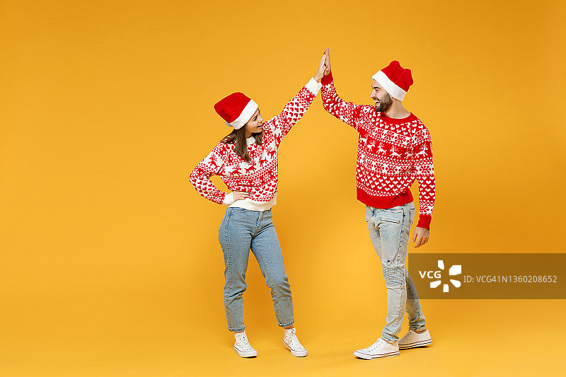 长得欢快的年轻圣诞老人夫妇朋友，男人女人在毛衣，圣诞帽给击掌看彼此孤立在黄色的背景。祝新年欢度佳节理念。图片素材