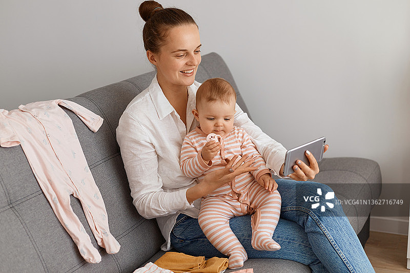 室内拍摄的一名梳着发髻、穿着白色衬衫和牛仔裤的女性，手拿智能手机，抱着婴儿坐在沙发上，在社交网络上直播。图片素材