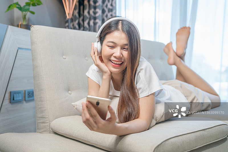 漂亮的年轻女子戴着耳机在沙发上放松。女性用智能手机听音乐。冷静休闲的概念图片素材