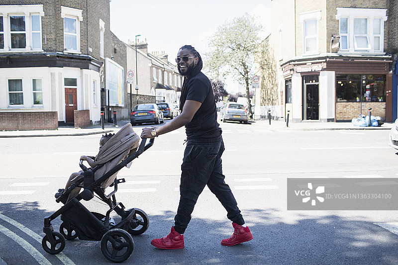 快乐的父亲推着婴儿车在阳光明媚的城市街道上蹒跚学步的儿子图片素材