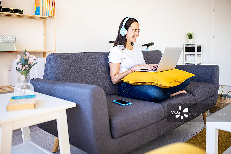 年轻的成年女性坐在家里的沙发上，一边用笔记本电脑工作，一边戴着耳机听音乐。图片素材