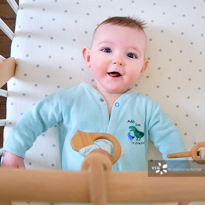 快乐的婴儿宝宝正躺在床上玩着木制的挂玩具。一个六个月大、穿着绿松石色衣服、面带微笑的滑稽孩子躺在婴儿床上图片素材