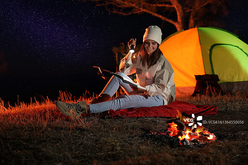 一个年轻的女人拿着手电筒在篝火旁看书。露营的季节图片素材