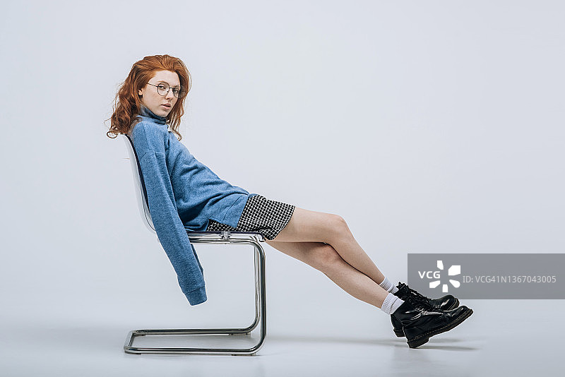 一个红头发的年轻女孩坐在工作室白色背景的椅子上摆姿势。图片素材