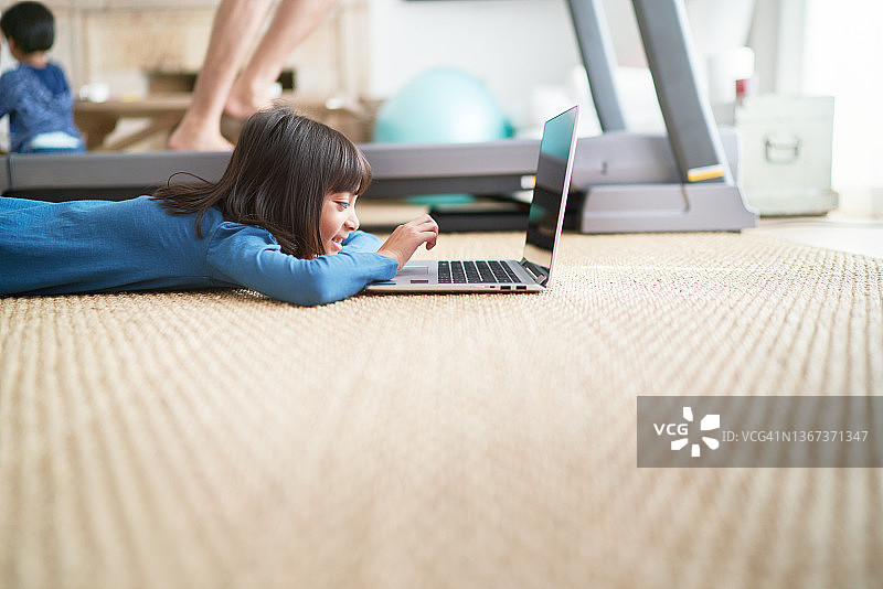 女孩在客厅地板上用笔记本电脑，旁边的爸爸在跑步机上图片素材