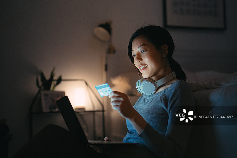 一名年轻的亚洲女性在深夜坐在卧室里，用笔记本电脑上网和购物，用信用卡进行移动支付。安全便捷的网上购物体验。科技智能银行图片素材