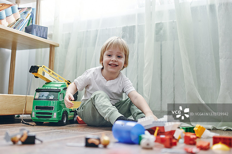 一个3岁的小男孩，一头金发，身穿白色t恤，面带微笑地坐在木地板上玩着五颜六色的方块图片素材