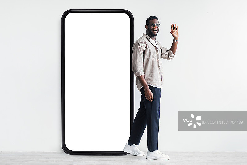 一个快乐的黑人站在白色屏幕的巨型智能手机旁，对着相机挥手问好图片素材
