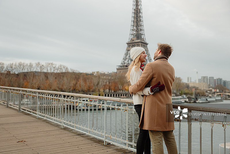 一对成年夫妇站在塞纳河上的桥上，拥抱着对方，看着对方，背景是埃菲尔铁塔图片素材