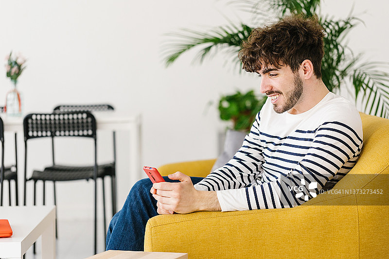 一个微笑的男人坐在家里的沙发上玩手机。图片素材