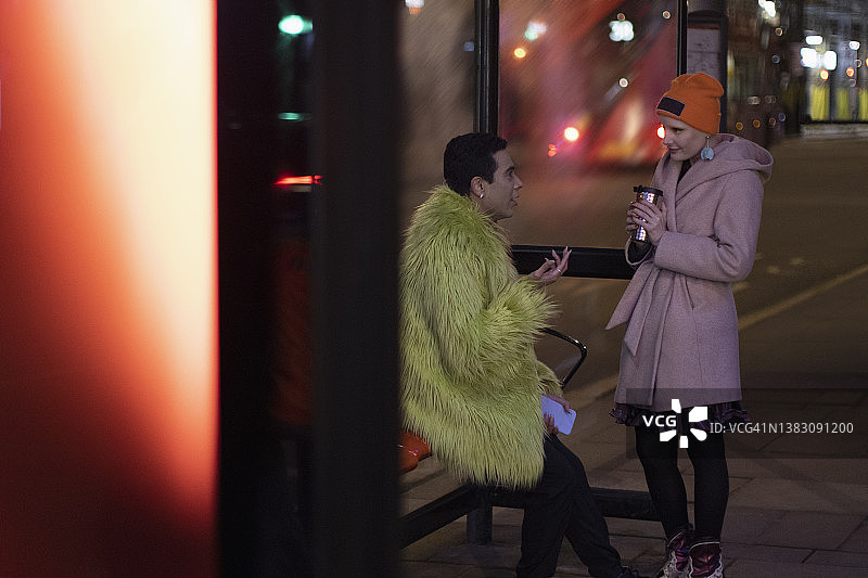 一对时髦的夫妇晚上在城市的公交车站喝着咖啡聊天图片素材