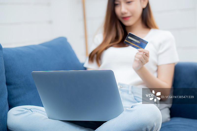 年轻的亚洲女人坐在沙发上使用笔记本电脑购物网上信用卡购买到互联网，快乐的女孩支付与电子商务在沙发上，购买和支付，商业理念。图片素材
